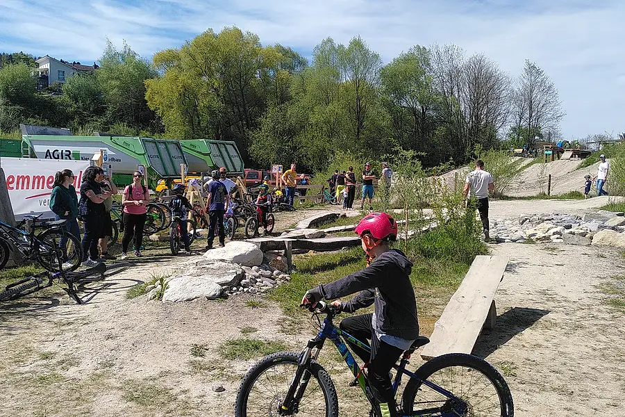 Ein junger Mountainbiker mit rotem Helm fährt an, dahinter Besuchergruppen und abgestellte Bikes.
