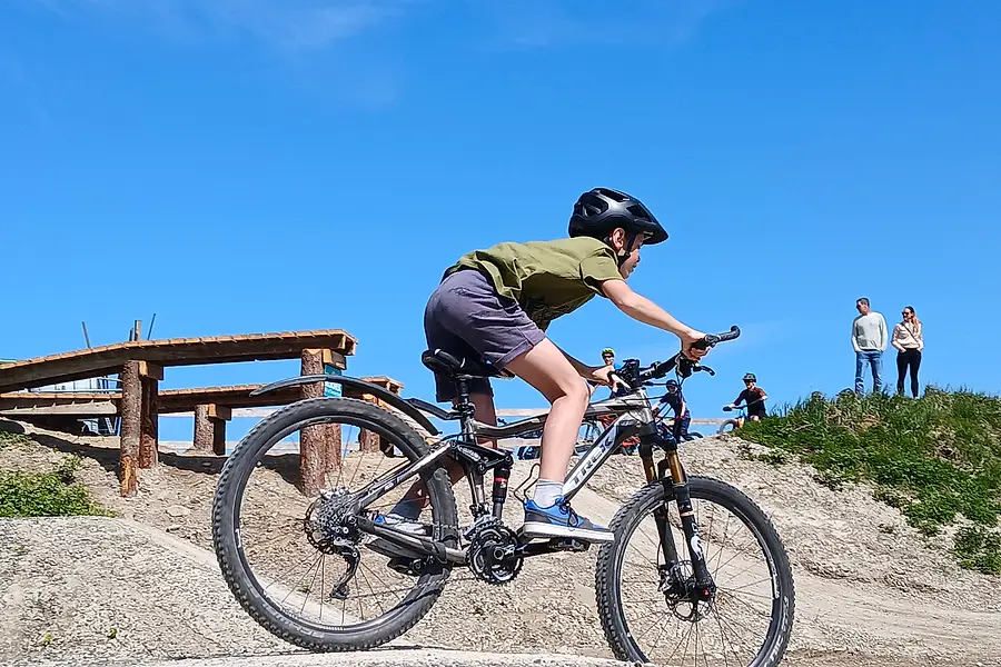 Ein jugendlicher Fahrer im Profil über die Kuppe, dahinter das Holz-Element und zuschauende Erwachsene.