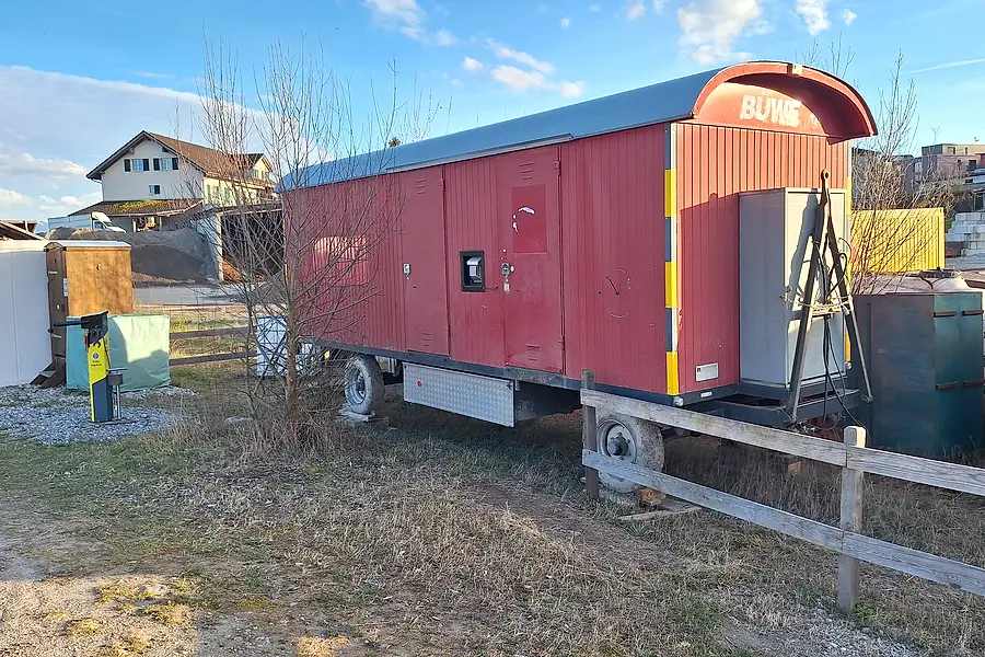 Der rote Bauwagen in voller Seitenansicht auf dem Areal, links der weisse Container.