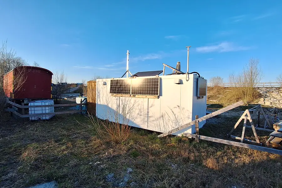 Der Container mit Solarpanel und Kaminrohr, der seit Anfang dabei ist, davor Holzböcke und Bretter.