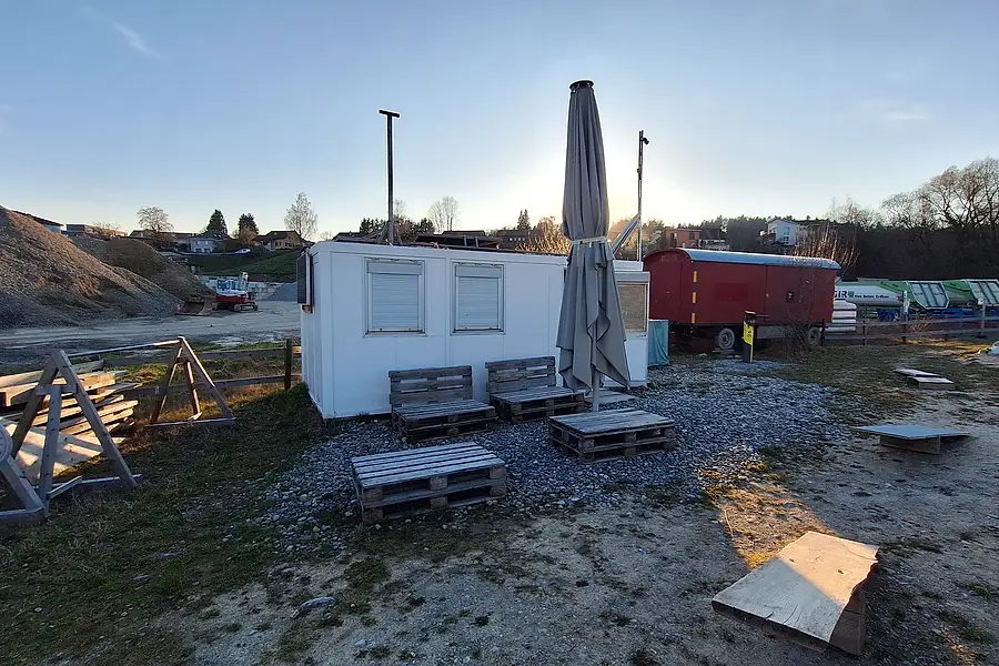 Weitwinkelblick über den Platz mit Wohncontainer, Bänken und Palettenboden, rechts der rote Bauwagen.