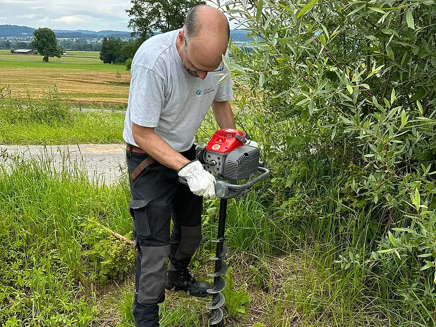 Ein Helfer bohrt mit dem Motor-Erdbohrer ein Pfostenloch am Rand der Fläche, dahinter die Felder Richtung Dorf.