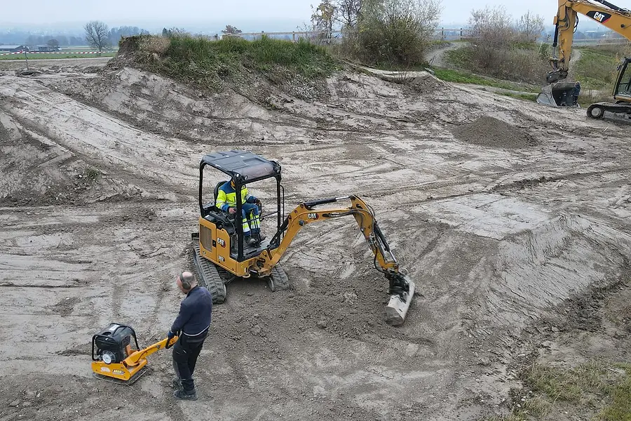 Ein Minibagger formt ein Landefeld, daneben verdichtet ein Helfer mit der Rüttelplatte.