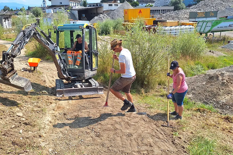 Ein Minibagger hebt das Terrain aus, zwei Helfer stecken mit Rechen und Messlatte die Höhe der neuen Linie ab.